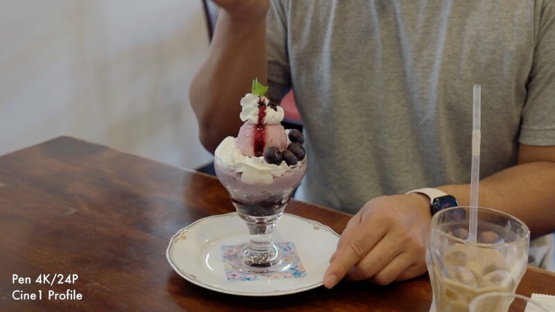 A person sits at a wooden table next to a blueberry parfait in a glass dish and a glass of iced coffee.