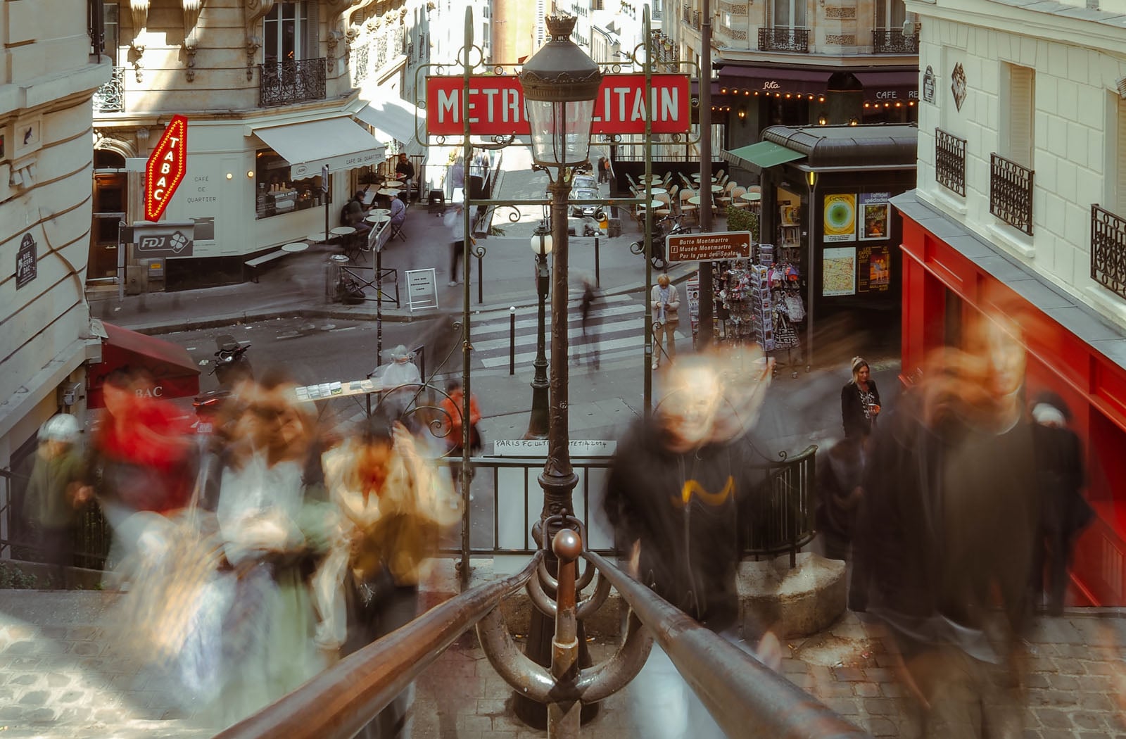 A high-angle view of a Parisian street corner with a Metro entrance, featuring blurred pedestrians moving down a staircase in the foreground.