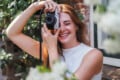 A smiling woman with red hair and freckles holds a silver camera up to her face, partially obscuring one eye.