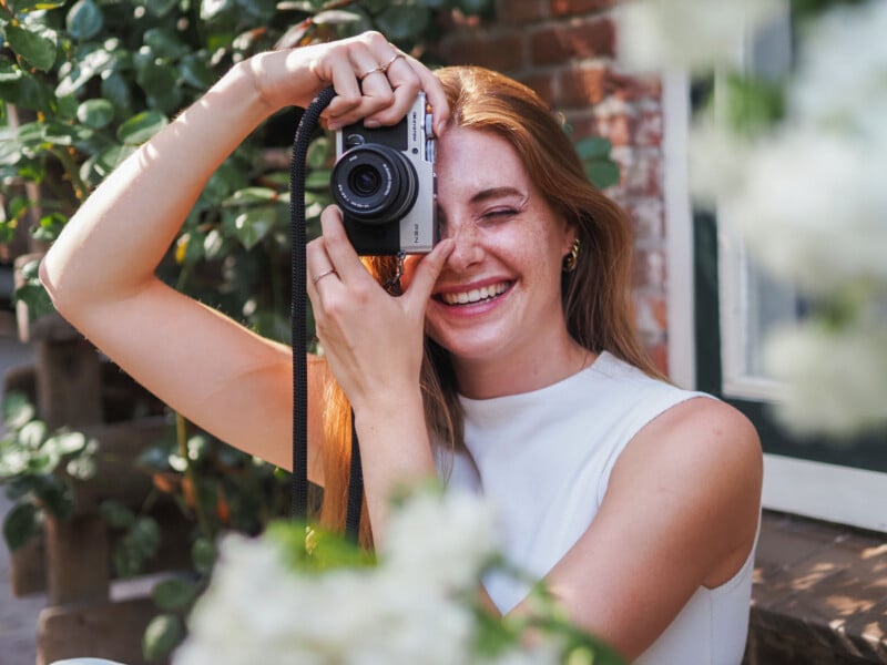 A smiling woman with red hair and freckles holds a silver camera up to her face, partially obscuring one eye.