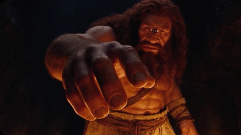 A large, muscular cyclops with long brown hair and a beard reaches his hand toward the viewer in a dimly lit setting.