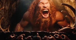 A giant, red-bearded cyclops roars at a group of armored warriors standing in a dark cave.