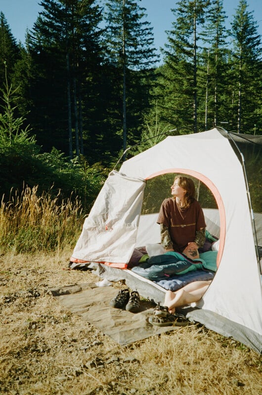A person sits inside a tent in a grassy clearing surrounded by a forest, with a pair of sandals resting on a ground cloth outside the entrance.