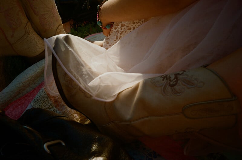 A person wearing a white lace dress and cowboy boots sits on a patterned quilt next to a black leather bag.