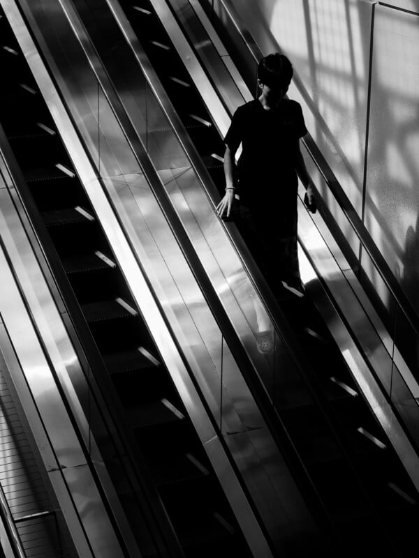 A person in a black t-shirt descends a metal escalator, captured in a high-contrast black and white shot.