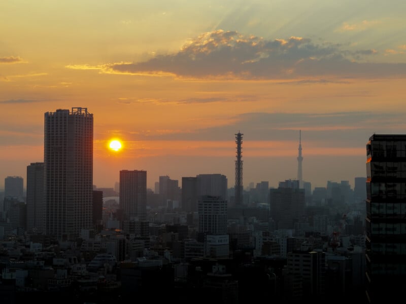 A sunset over a dense city skyline with a tall communications tower and the Tokyo Skytree visible in the distance.