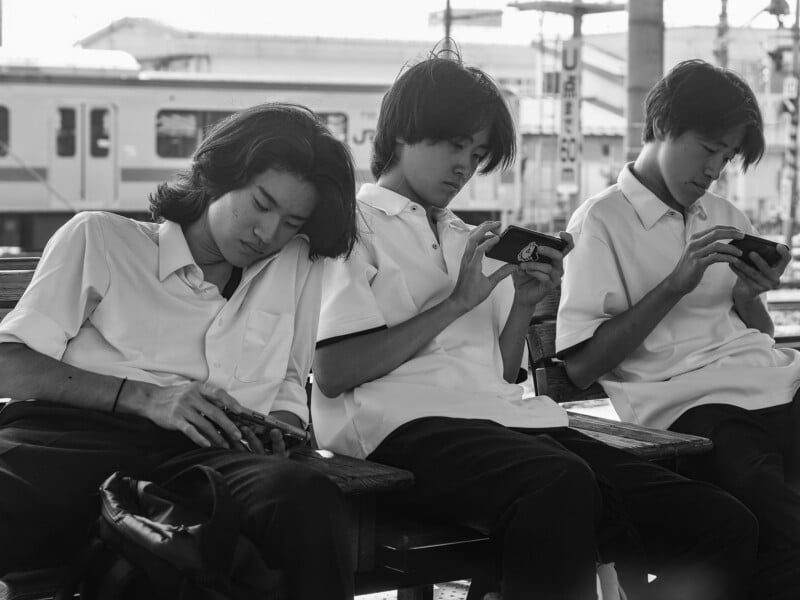 Three young men in white collared shirts sit on a wooden bench at a train station, each looking down at a smartphone.