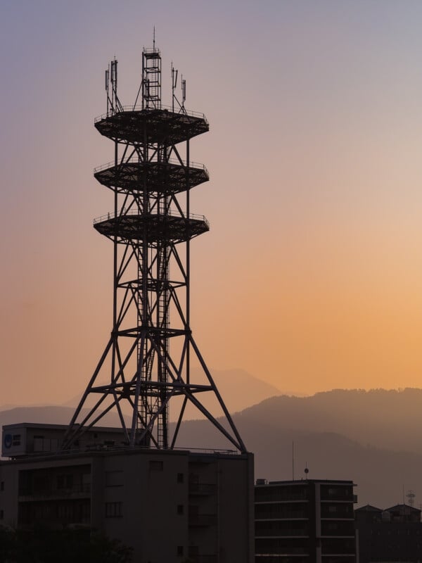 A tall metal telecommunications tower stands silhouetted against a sunset sky, rising above a building with mountains in the distance.