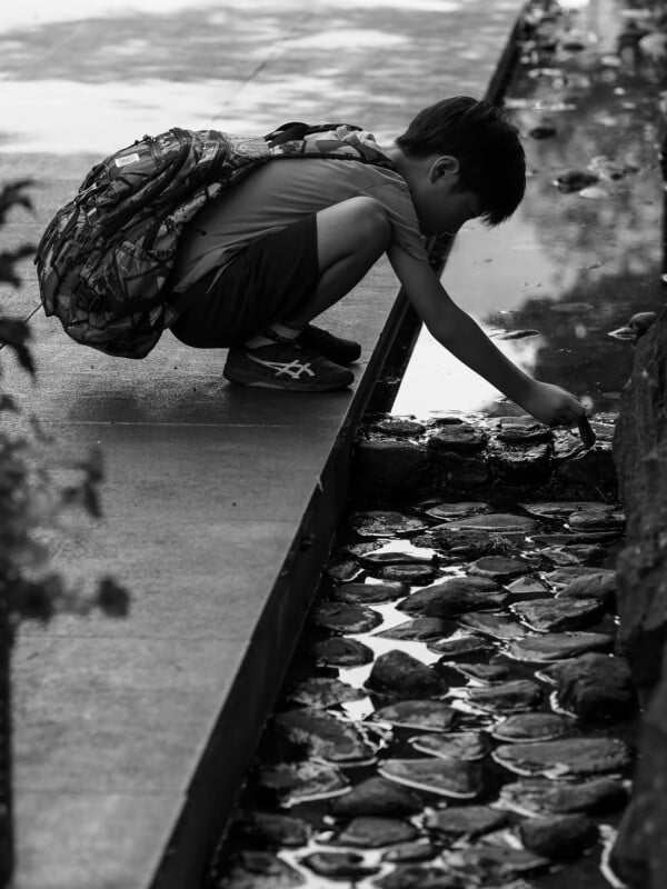 A young boy with a backpack crouches on a stone ledge, reaching down to touch the water in a shallow, rocky stream.