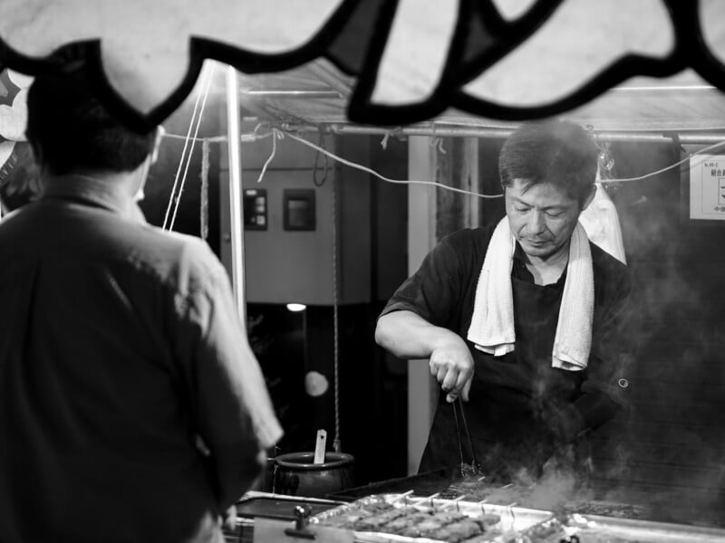 A man with a towel around his neck uses tongs to grill skewers at a street food stall while a customer stands in the foreground.