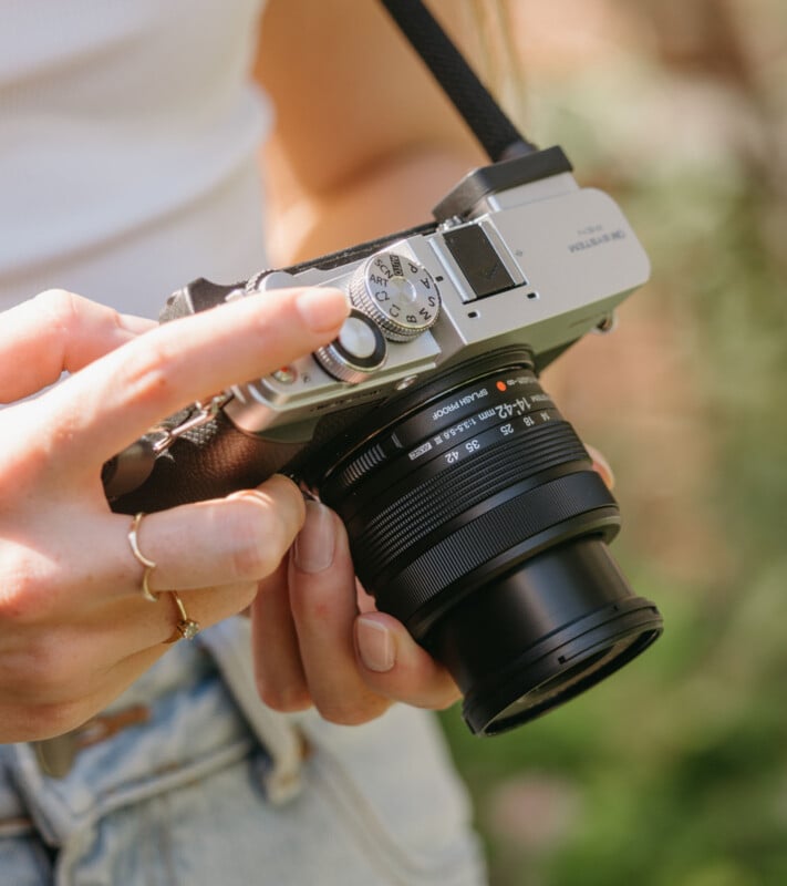 A person's hands hold a silver and black digital camera with a zoom lens, with their finger resting on the mode dial.