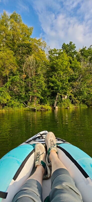 A first-person view from a blue and white inflatable kayak, showing a person's legs and light-colored water shoes against a backdrop of green trees and a blue sky.