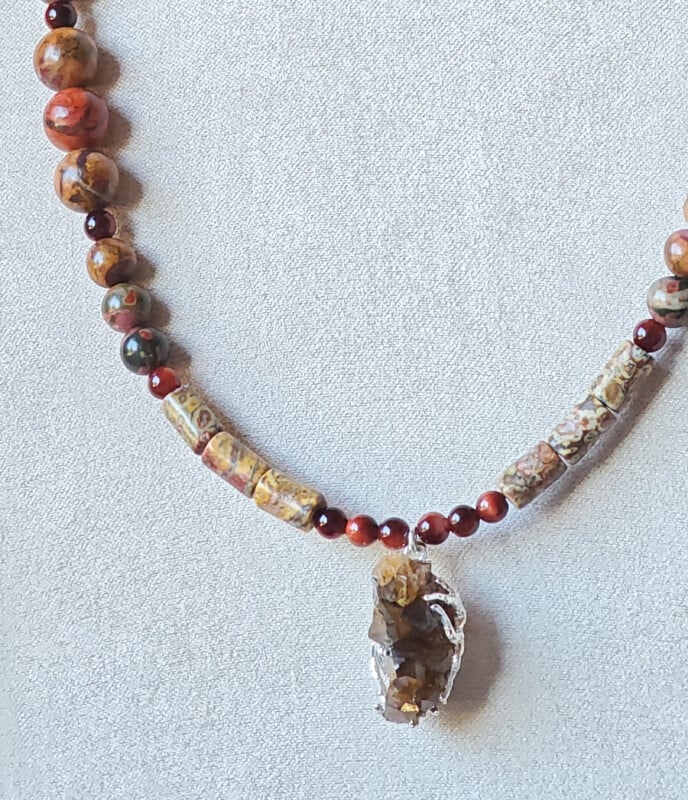 A beaded necklace featuring round and cylindrical jasper stones in earth tones, accented with small red beads and a central, wire-wrapped raw crystal pendant.