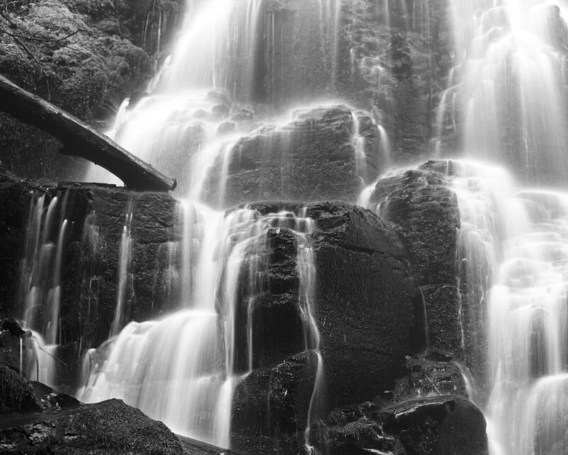 A black and white long-exposure shot of a waterfall cascading over dark, mossy rock formations with a fallen log on the left.