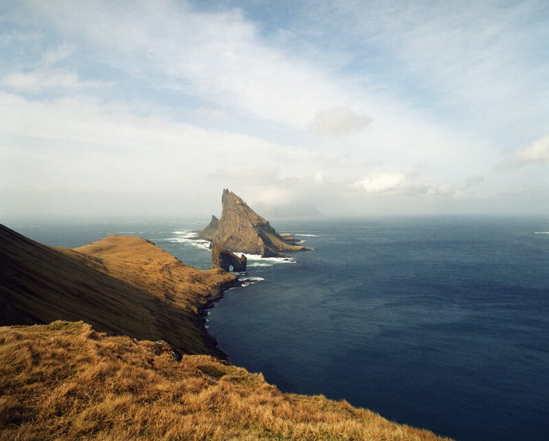 A grassy cliffside overlooks a deep blue ocean with a jagged, pointed rock formation rising from the water in the distance.