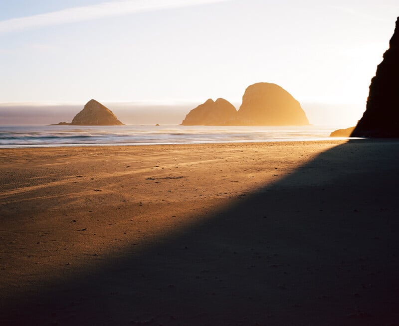 A sandy beach at sunset with large rock formations in the distance and a long shadow cast across the foreground.