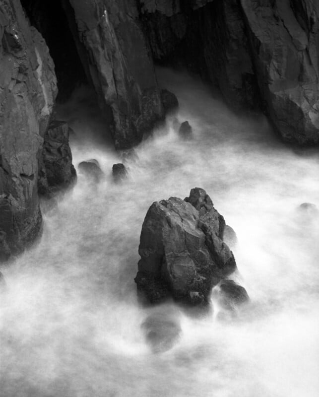 A long-exposure, black-and-white view of dark, jagged rocks surrounded by swirling, blurred ocean surf.