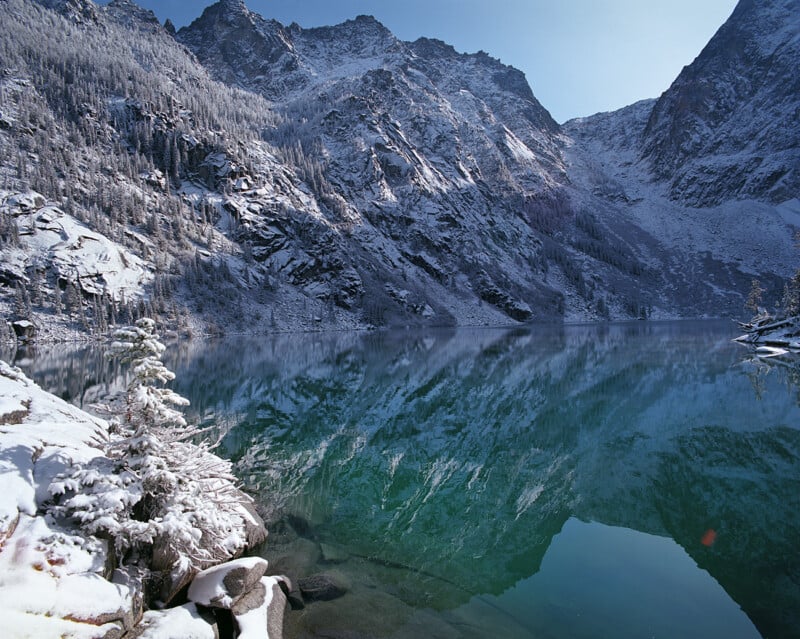 A snow-covered mountain range reflects into the calm, turquoise water of an alpine lake, with a small, frosted evergreen in the foreground.