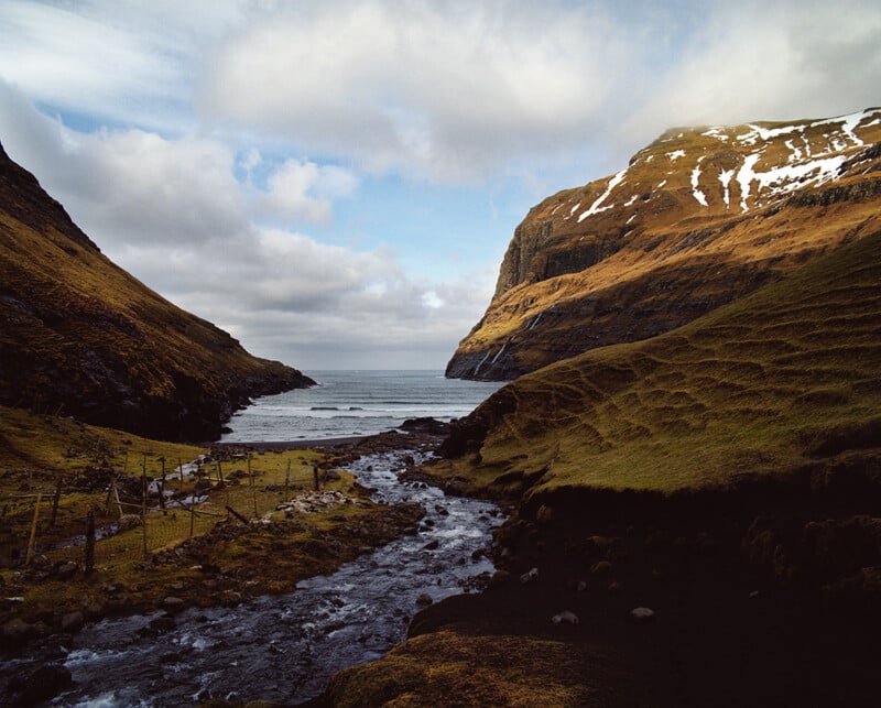 A stream flows through a grassy valley between two large, steep hills toward a dark sand beach and the ocean.