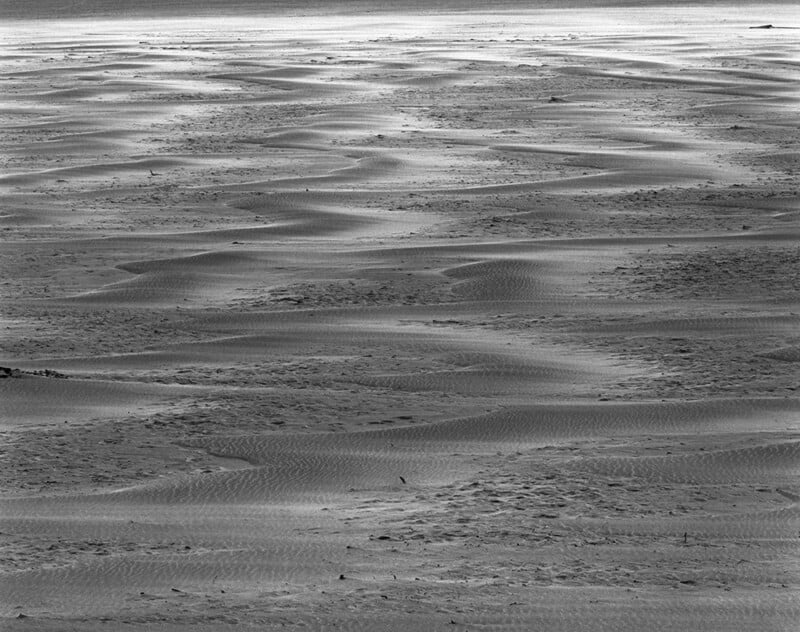 A black and white landscape showing rippled sand patterns stretching across a beach toward a bright horizon.