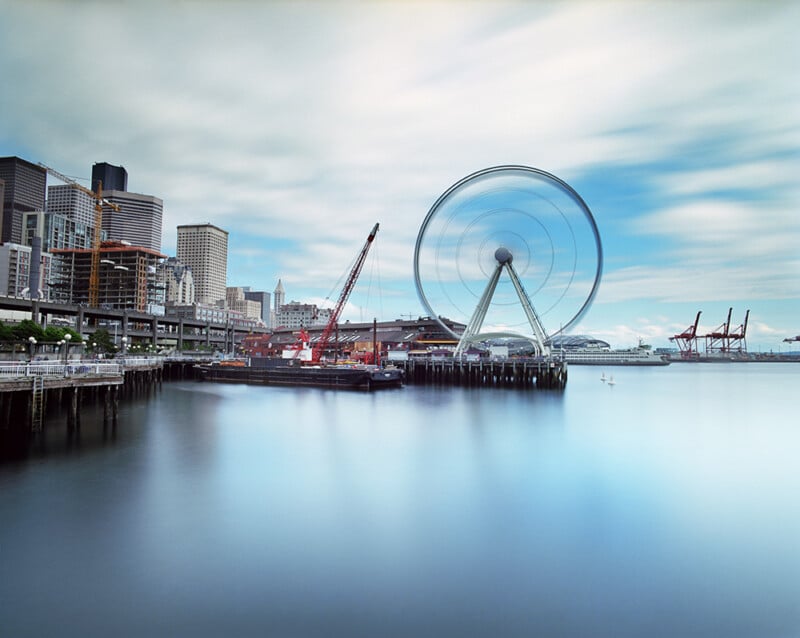 A long-exposure view of the Seattle waterfront featuring the Great Wheel in motion, a construction crane, and a ferry on the water.