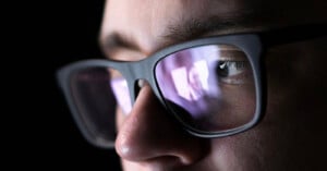 A close-up, side-profile view of a person wearing black-rimmed glasses, with a bright reflection visible on the lenses.