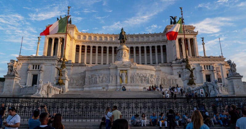 The Altare della Patria in Rome features a grand white marble monument with a central equestrian statue, flanked by two Italian flags, with people gathered on the steps below.