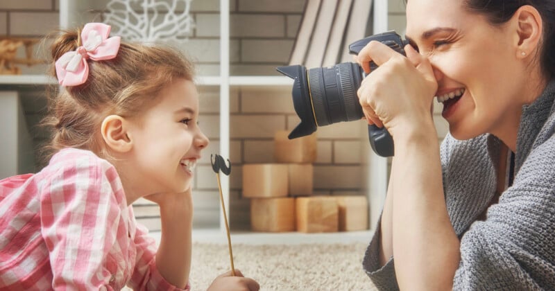 A woman smiles while taking a picture of a young girl holding a mustache prop on a stick.
