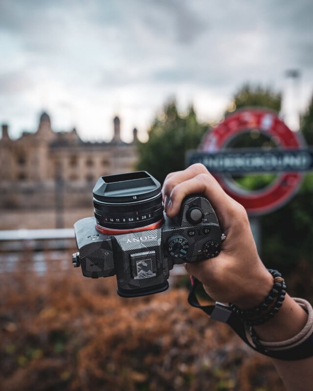 A person holds a camera with a patterned skin and a lens hood in front of a blurred London Underground sign and a building.
