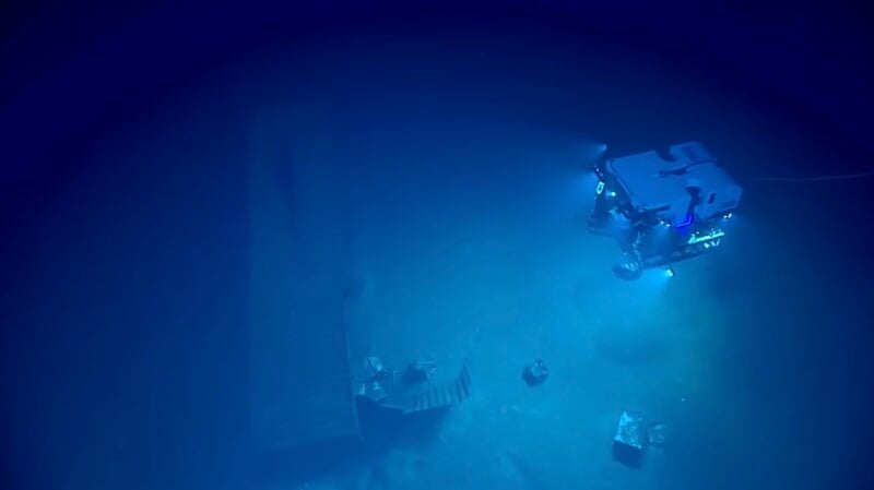 A remotely operated underwater vehicle illuminates the dark, deep-sea floor, where a wooden structure and scattered debris rest on the sediment.