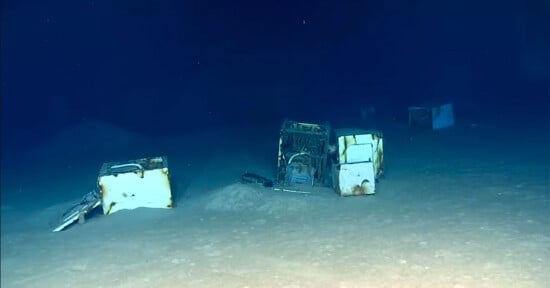 Several discarded white appliances, including washing machines, sit partially buried in the sandy sediment on the dark ocean floor.