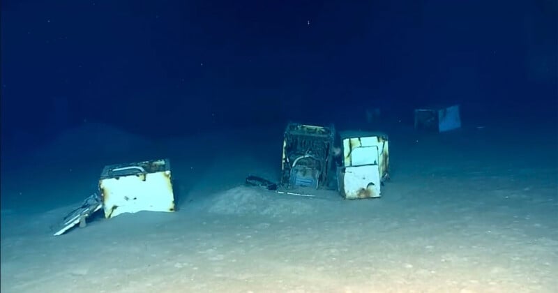 Several discarded white appliances, including washing machines, sit partially buried in the sandy sediment on the dark ocean floor.