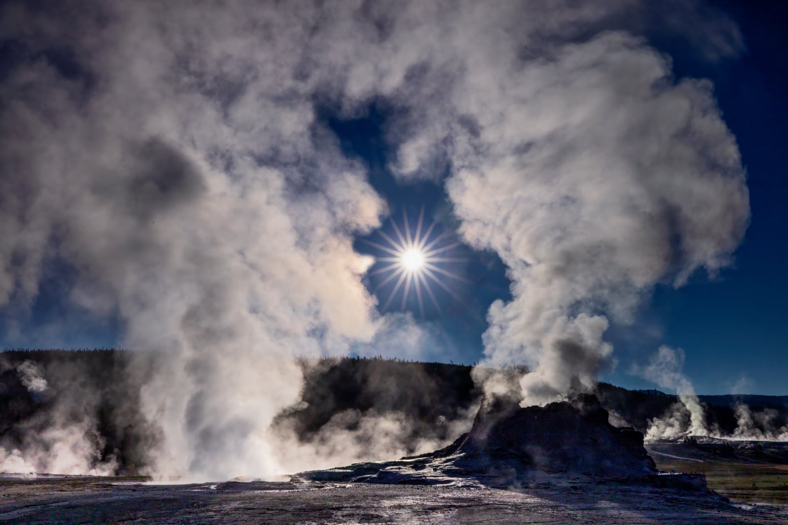 Sunlight shines through a gap in thick steam rising from a rocky, geothermal landscape under a deep blue sky.