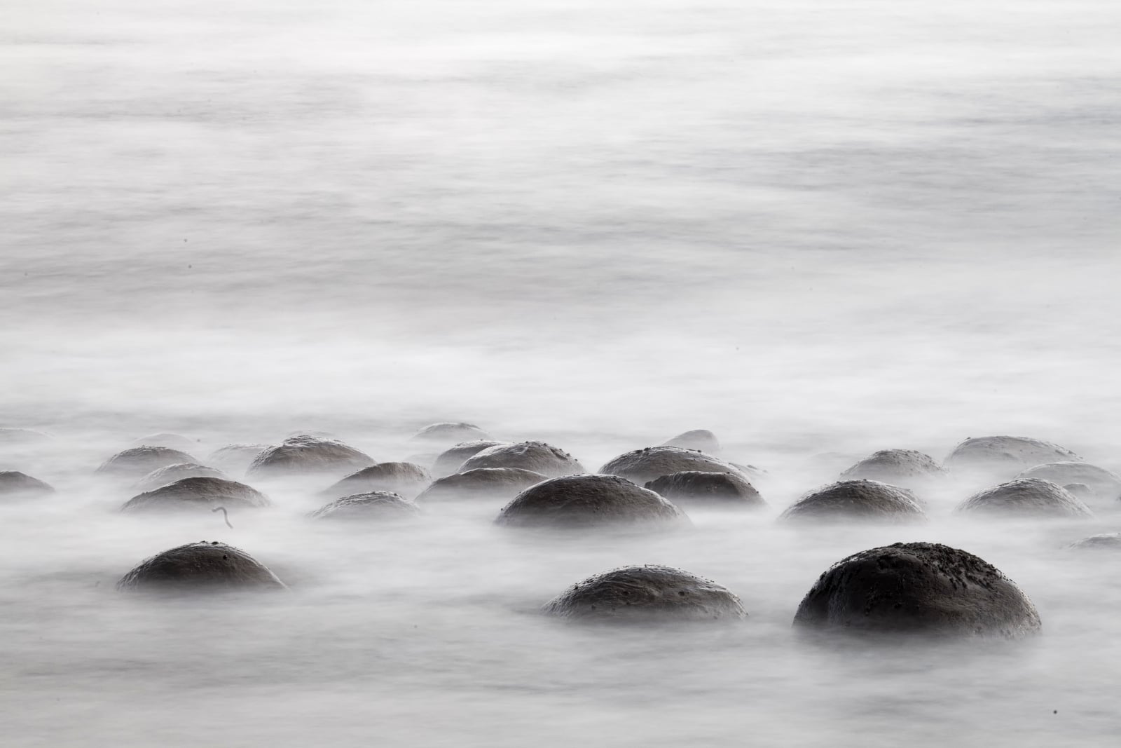 A cluster of smooth, dark boulders sits partially submerged in a calm, misty body of water with a long-exposure effect.