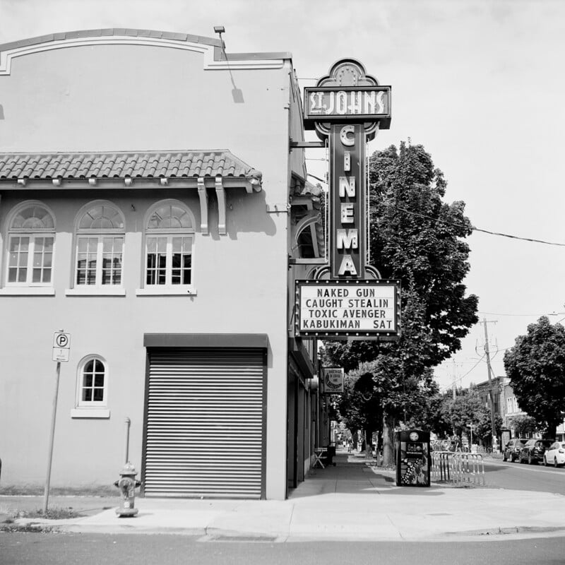 A black-and-white street-level view of the St. Johns Cinema building, featuring a vertical marquee sign listing movie titles and a closed metal storefront shutter.