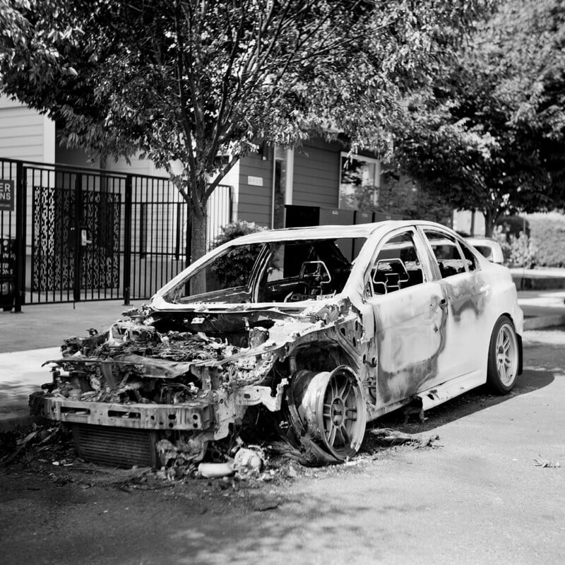 A severely fire-damaged sedan sits abandoned on a city street, missing its front bumper, hood, and front tire.