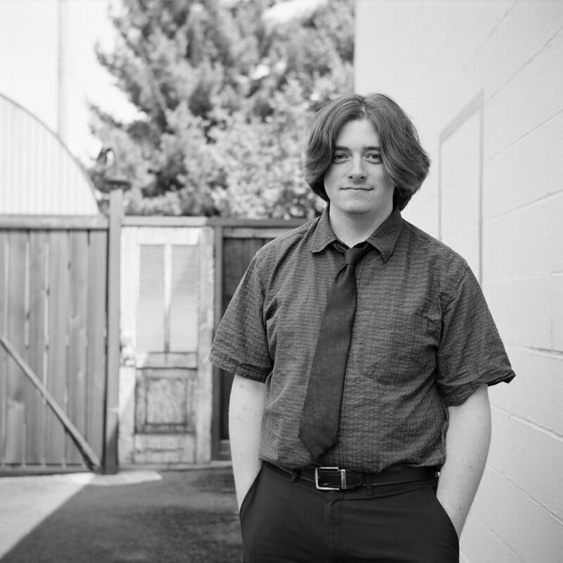 A young person with shoulder-length hair stands outdoors wearing a short-sleeved button-down shirt, a tie, and trousers with hands in their pockets.