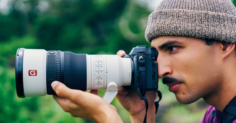 A person wearing a beanie looks through the viewfinder of a large white telephoto camera lens held with both hands.