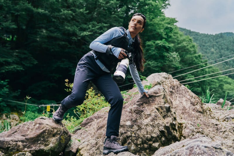 A woman wearing a light blue long-sleeved shirt, dark pants, and hiking boots climbs over large rocks while holding a camera with a long lens.