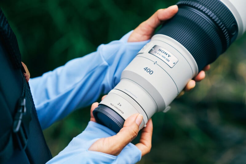 A person wearing a light blue long-sleeved shirt holds a white Sony FE 4.5/400 GM lens with a 2x teleconverter attached.