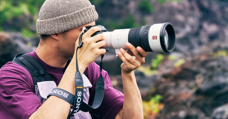 A person wearing a beanie and a purple shirt holds a camera with a large white telephoto lens to their eye.