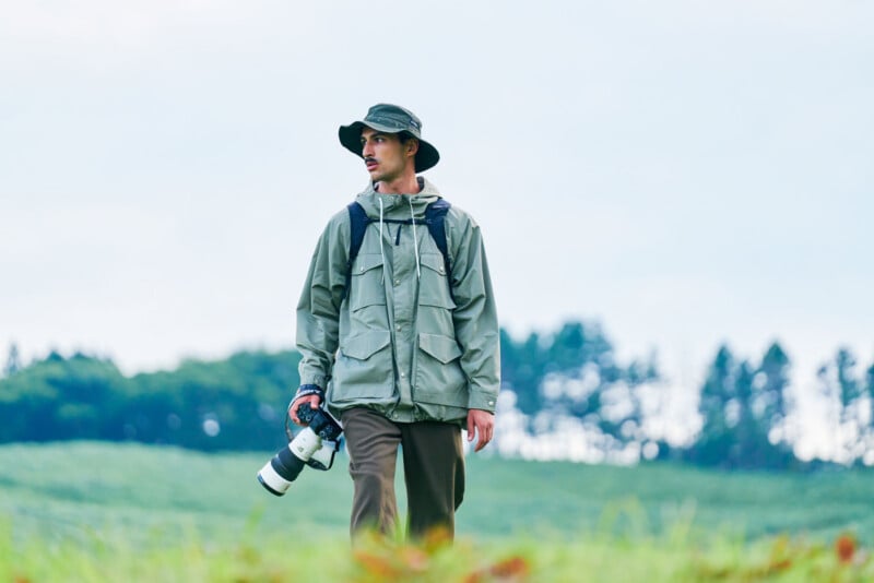 A man with a mustache wearing a bucket hat and a light green jacket walks through a grassy field while holding a camera with a large lens.