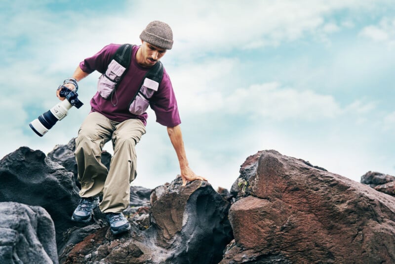 A man wearing a beanie, purple shirt, and utility vest climbs over dark volcanic rocks while holding a camera with a long lens.