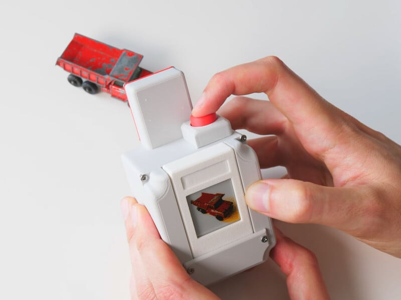 A person holds a white, 3D-printed handheld device with a small screen displaying a red dump truck, while a matching toy truck sits on the white surface nearby.