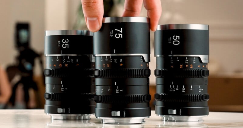 A hand holds the middle lens of three black cinema lenses labeled 35, 75, and 50, standing upright on a table.