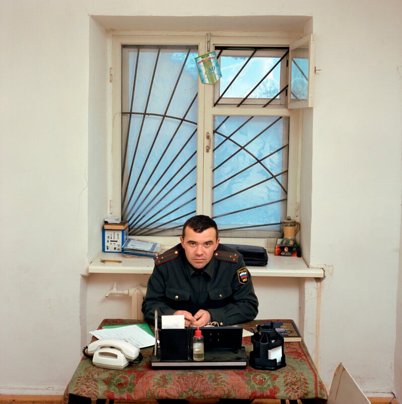 A man in a Russian military uniform sits at a desk in front of a barred window.