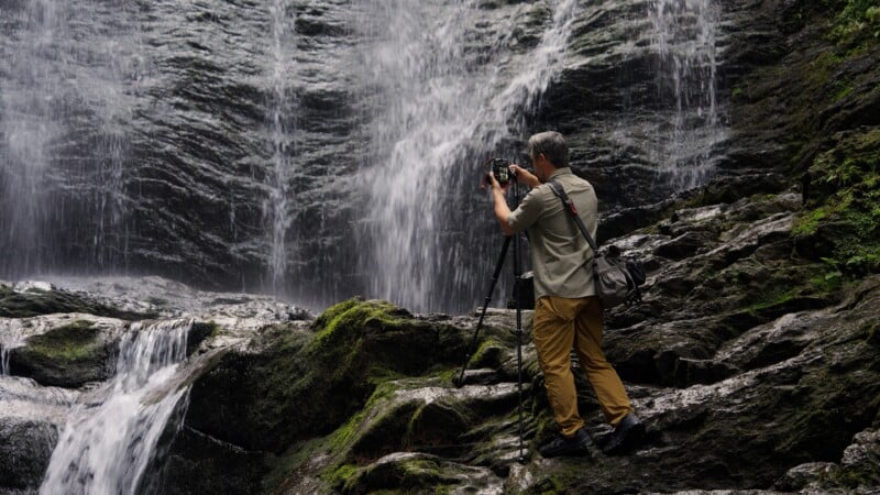 A man with gray hair stands on mossy rocks, using a camera on a tripod to photograph a waterfall.