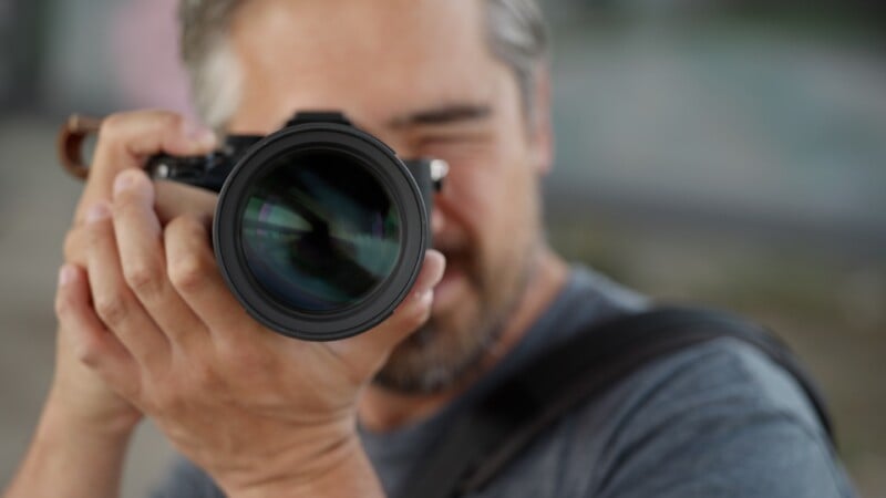 A man with gray hair holds a camera up to his eye, focusing the lens toward the viewer.