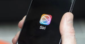 A hand holds a smartphone displaying a colorful, infinity-shaped icon above the word "Siri" on a black background.