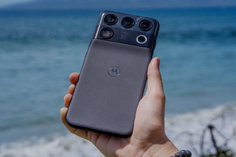 A hand holds a dark gray smartphone with a textured back and a prominent camera module against a blurred ocean background.